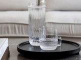 En vandkaraffel i riflet glas og to matchende glas står på en sort, rund bakke, der er placeret på et bord med en lys sofa i baggrunden.
