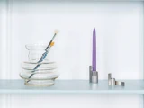 En klar, bølget glasvase med pensler indeni står ved siden af et højt lilla lys i en moderne sølvholder på en lyseblå hylde.