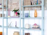 Lyseblå hylder med bøger, stearinlys, planter, garn, glas og pyntegenstande mod en hvid panel- og murstensvæg.