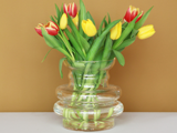 En klar glasvase med gule og røde tulipaner står på en hvid flade mod en lysebrun baggrund.