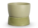 En beige keramikpotte står oven på en omvendt, lidt mørkere grøn underkop, begge med en glat, mat finish på en hvid baggrund.