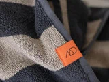 Nærbillede af et stribet håndklæde med marineblå og lysegrå bånd og et orange mærke med et minimalistisk sort AD-logo.