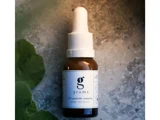 En brun glasdråbeflaske med påskriften grums HA-peptide complex under eye serum står på en struktureret overflade med et grønt blad i hjørnet.