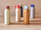 Fem farverige vandflasker med matchende hætter og håndtag er arrangeret på en lys træoverflade mod en beige træbaggrund.
