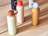 Fire farverige vandflasker med skruelåg og fastgjorte håndtag er arrangeret på en lys træoverflade. Flaskerne er i beige, solbrune, brune og gule nuancer.