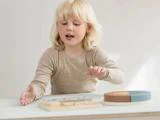 Et lille barn med blondt hår leger med et træpuslespil ved et hvidt bord og rækker interesseret og fokuseret frem.