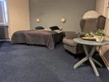 Et hyggeligt soveværelse med en dobbeltseng, gråt sengetøj, en lænestol og et rundt sidebord med en plante og en bakke på, sat op mod blåt gulvtæppe og blød belysning.