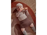 En baby iført en strikket hue, en blomstret bluse, brune shorts og grå sokker ligger på et tæppe og holder om og tygger på et blødt legetøj.