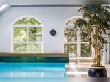 Indendørs swimmingpool med klart blåt vand, store buede vinduer med udsigt til grønne træer udenfor, et ur på væggen og en potteplante i højre side.