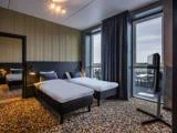 Moderne hotelværelse med to enkeltsenge, et stort vindue med udsigt over byen, mørke gardiner, et tv på en stander, et vægspejl og et mønstret gulvtæppe.