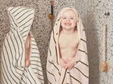 To små børn indhyllet i stribede håndklæder står i et flisebadeværelse. Det ene barn smiler til kameraet, mens det andet for det meste er dækket af sit håndklæde. En badebørste af træ hænger på væggen