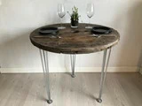 Et rundt træbord med hårnåleben i metal, dækket til to med sorte tallerkener, sølvtøj, vinglas og en lille potteplante i midten.