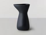 En matsort keramisk kande med et asymmetrisk, moderne design står på en lysegrå overflade mod en lys baggrund.
