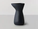 En matsort keramisk kande med et asymmetrisk, moderne design står på en lysegrå overflade mod en lys baggrund.