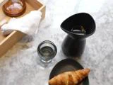 En croissant på en sort tallerken, en lille sort kande, et glas vand og en træbakke med en hvid serviet på en marmoroverflade.