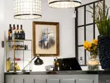 En moderne receptionsdisk med flasker på hylder, en skrivebordslampe, gule blomster og et indrammet portræt af en ældre mand på væggen. To store pendler hænger fra loftet.
