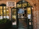Indgangen til Station Cafe med åbne dobbeltdøre, murstensvægge, store vinduer og et skilt over døren. Inde i lokalet kan man se borde og planter.