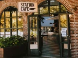 Indgangen til en café med murstensvægge, store vinduer, åbne dobbeltdøre og et skilt med teksten »Station Cafe«. Udenfor ses grønne omgivelser, og inde i lokalet ses en disk.