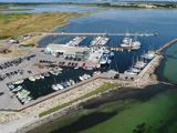 Luftfoto af en marina med dokker, både og yachts, omgivet af vand og græsarealer, med biler parkeret i nærheden.