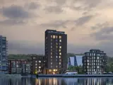 Høje moderne boligblokke med store vinduer spejler sig i roligt vand i skumringen, med en bus parkeret i nærheden og træer i baggrunden under en overskyet himmel.