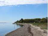 En rolig stenstrand buer sig langs det stille, blå vand, omgivet af grønne træer og et par små huse under en delvis overskyet himmel.