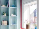 Lyseblå hylder med forskellige ting, bl.a. planter, bøger og tallerkener, ved siden af et solbeskinnet vindue med blomster i vaser og udsigt til huse udenfor.