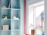 Lyseblå hylder med forskellige ting, bl.a. planter, bøger og tallerkener, ved siden af et solbeskinnet vindue med blomster i vaser og udsigt til huse udenfor.