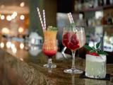 Tre farverige cocktails med stribede sugerør og pynt står på en bardisk med en sløret baggrund af flasker og varm belysning.