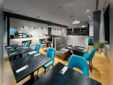 Moderne spiseplads med mørke træborde, blå stole, grå siddepladser og pendellamper. Buffetopstilling og kaffemaskiner ses i baggrunden; potteplante i hjørnet.