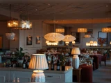 En varmt oplyst restaurant med elegante hængelamper, dækkede borde, vinflasker og indrammet kunst på væggene, som skaber en hyggelig og indbydende atmosfære.