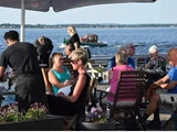 Folk spiser udendørs på en restaurant ved vandet på en solrig dag med en båd og roligt vand i baggrunden.