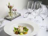 En anrettet gourmetret med fisk, grøntsager og krydderurter på et hvidt bord, omgivet af tomme vinglas og en blomsterdekoration.