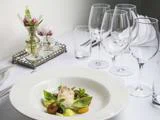 En gourmetret med fisk, grøntsager og krydderurter på en hvid tallerken, dækket på et elegant bord med tomme vinglas, vandglas, servietter og en lille blomsteropsats.