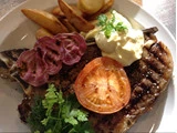 Grillet bøf med en skive tomat, kartoffelbåde, grillede lilla grøntsager, friske krydderurter og en klat cremet sauce på en hvid tallerken.