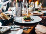 Nærbillede af et elegant spisebord dækket med vinglas, tallerkener med gourmetmad, brød og vand, hvor folk nyder et måltid i en varmt oplyst restaurant.
