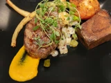 En gourmetret med en grillet bøf, gulerodspuré, ristet tomat, rodfrugter og en brunet rektangulær kartoffel på en sort tallerken, pyntet med friske krydderurter.
