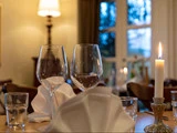 Nærbillede af et spisebord dækket med tomme vinglas, vandglas, foldede servietter og et tændt stearinlys i en hyggelig, svagt oplyst restaurant.