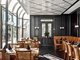 En stilfuld restaurant med træborde, kurvestole, brune læderstole, sorte servietter og store vinduer, der lukker naturligt lys ind. En potteplante står ved et centralt vindue.