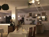 En hyggelig, varmt oplyst restaurant med træstole, opstillede borde, hængelamper og dekorative hylder bag et lille barområde.