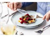 En person i blå skjorte bruger gaffel og kniv til at spise en farverig ret med grøntsager og krydderurter på en hvid tallerken ved et restaurantbord.