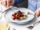 En person bruger kniv og gaffel til at spise en farverig gourmetret med grøntsager og mikrogrønt på en hvid tallerken.