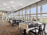 Moderne restaurant med store vinduer, opdækkede borde med sorte stole og hvide duge og udsigt til et græsareal og vandet udenfor.