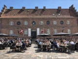 Folk, der sidder ved udendørsborde foran et stort murstenshotel med det danske flag.