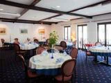 Elegant restaurantspisestue med runde borde dækket med hvide og blå duge, dækket med glas og servietter og omgivet af røde stole; store vinduer lukker naturligt lys ind.