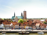Udsigt over en europæisk by med huse med røde tage, en stor kirke med to spir, et højt tårn og både, der ligger til kaj langs vandet under en klar himmel.