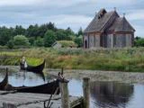 En person i en vikingebåd ror på en flod tæt på en trækirke og grønne marker, med træer og en overskyet himmel i baggrunden.