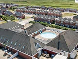 Luftfoto af et boligområde med rækker af røde murstenshuse og en central gårdhave med en cirkulær swimmingpool. Grønne marker strækker sig ud i baggrunden.