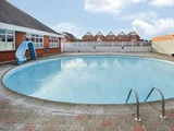 En rund udendørs swimmingpool med klart vand, metalstiger og en blå rutsjebane, omgivet af brolægning. I baggrunden ses rækker af huse under en delvist overskyet himmel.