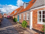 En brostensbelagt gade med charmerende murstenshuse med røde tegltage, hvide vinduer og grønne planter under en delvis overskyet himmel.