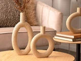 To teksturerede, ovale beige vaser med åben midte står på et lyst træbord i nærheden af stablede bøger og en moderne beige sofa i en hyggelig stue.